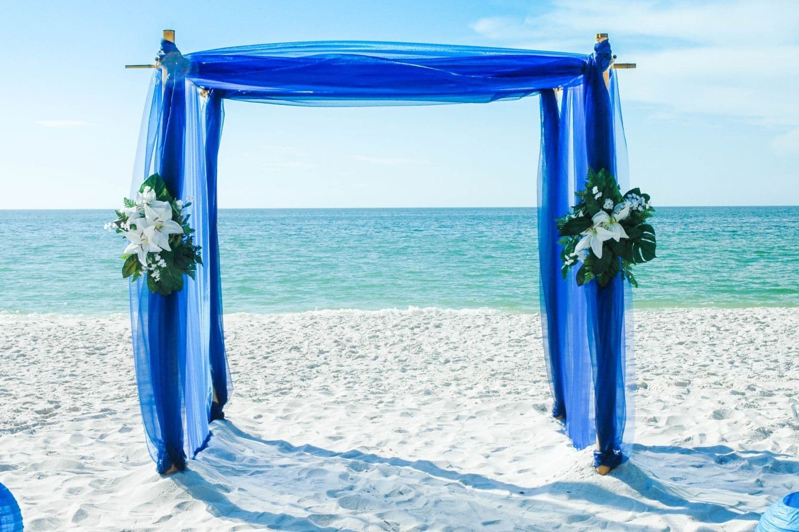 Blue draped wedding arch on a white sandy beach with ocean and sky in the background.