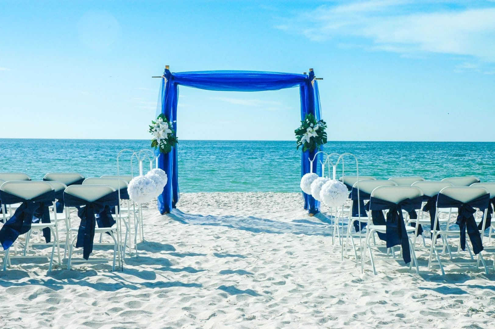 Wedding ceremony setup on a sandy beach with blue arch and ocean backdrop.