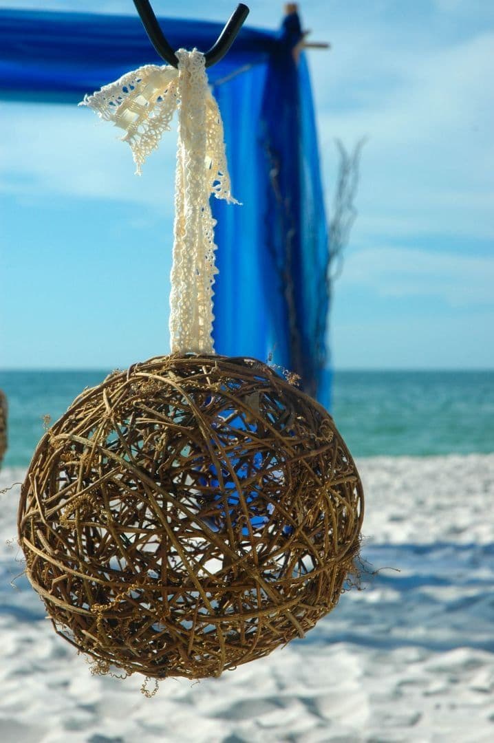 Woven sphere decoration, suspended by lace ribbon, against a blue beach wedding backdrop.