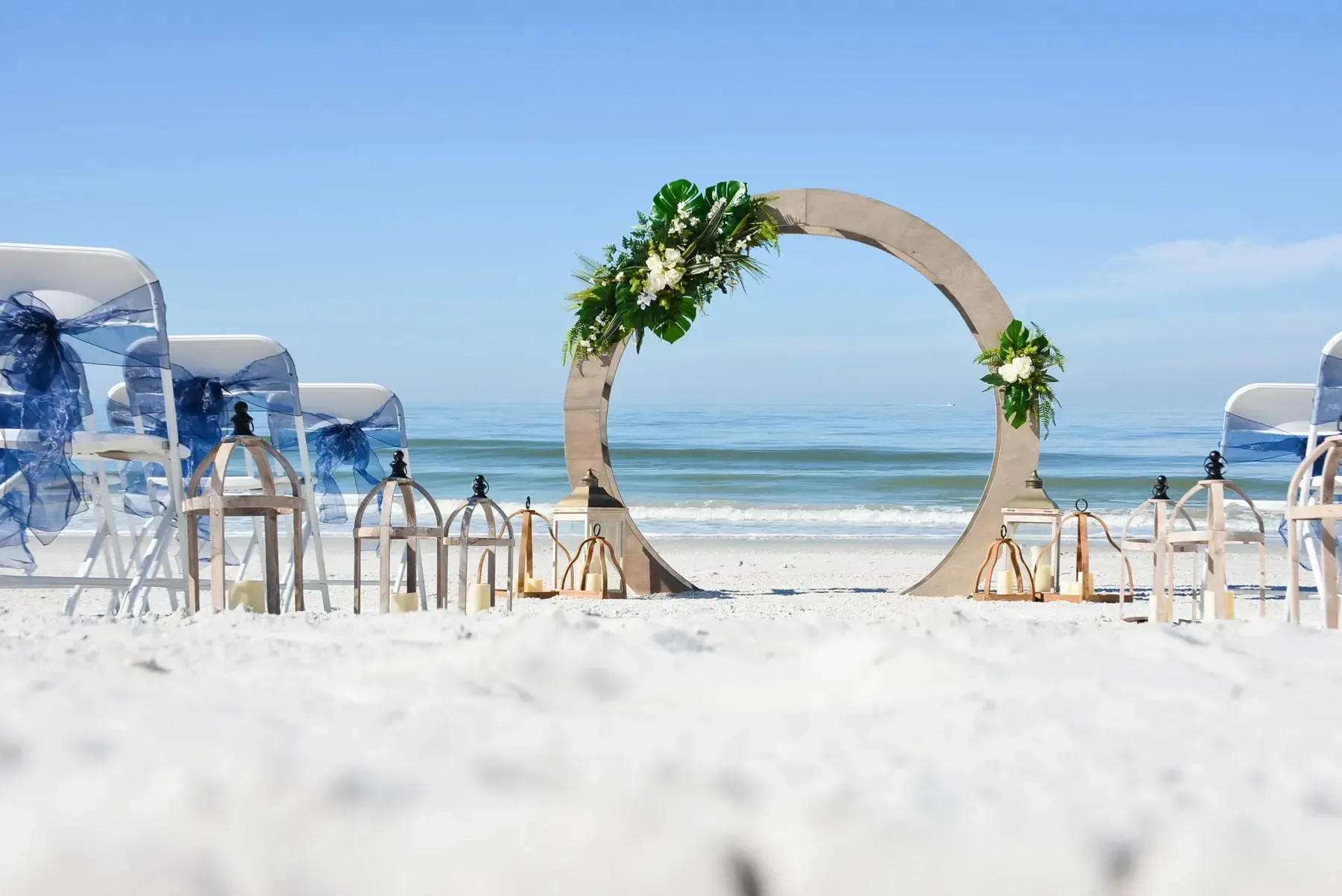 Beach wedding ceremony setup with an arch decorated with greenery and flowers, chairs facing the ocean.