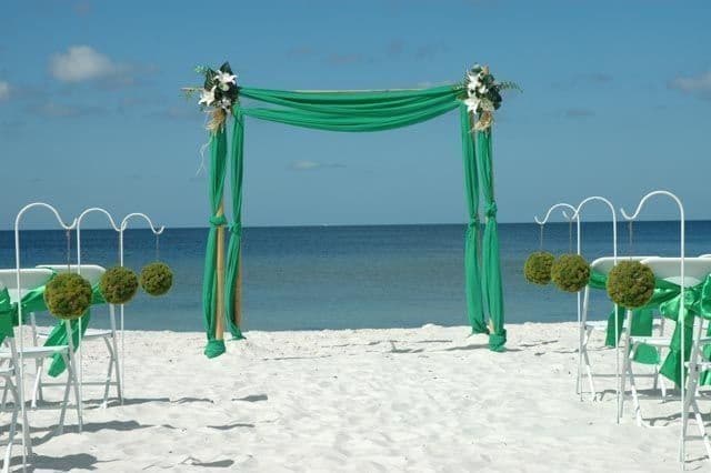Beach wedding setup with green draped arch, white chairs, and ocean backdrop.