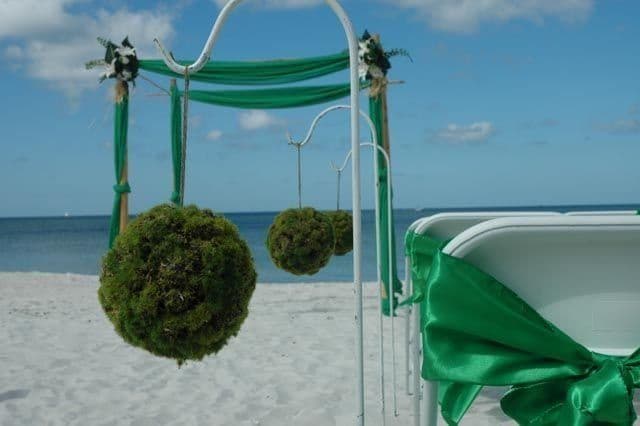 Wedding arch on a beach with green decorations, including hanging moss balls and chair bows.