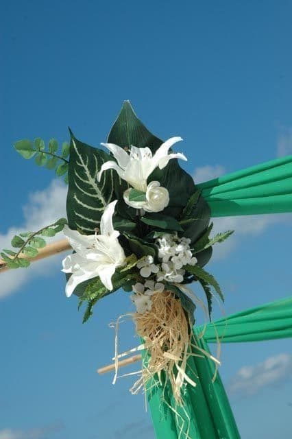 White lilies and greenery on a green fabric-draped structure against a bright blue sky.