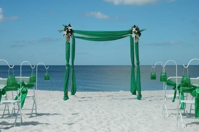 Wedding ceremony setup on a beach: white chairs, green draped arch, ocean, and blue sky.