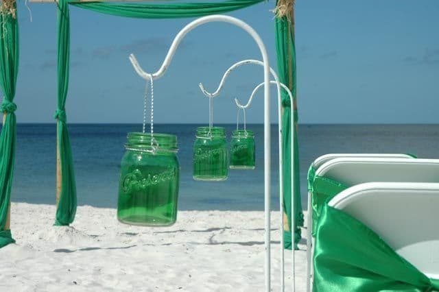 Beach wedding setup: Green mason jars hanging, white chairs with green sashes, turquoise ocean in background.