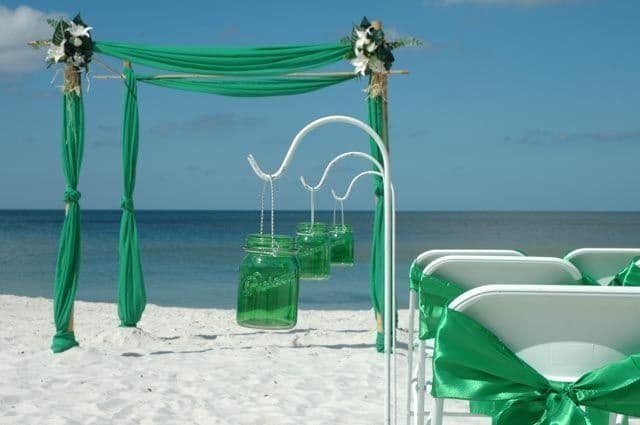 Beach wedding ceremony setup: emerald green draping, hanging jars, white chairs with green bows, ocean backdrop.