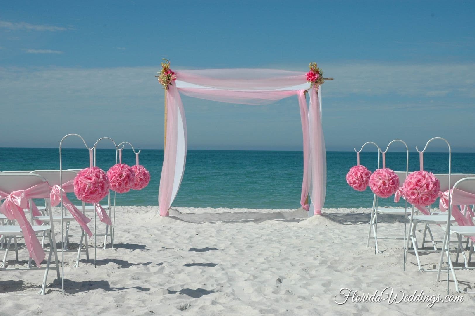 Beach wedding setup with pink chiffon arch, floral chair accents, and white chairs on a sandy beach.