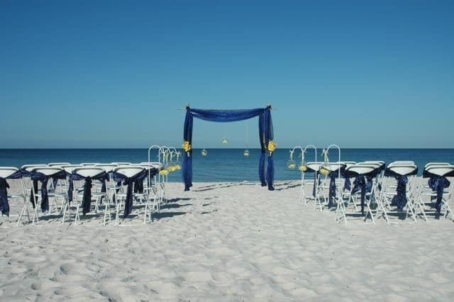 Beach wedding setup: white chairs with blue sashes face a blue draped arch with ocean and sky backdrop.