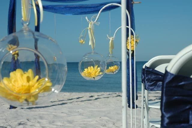 Wedding decorations on a beach: white arch with hanging glass orbs holding yellow flowers, blue fabric backdrop, ocean in the background.