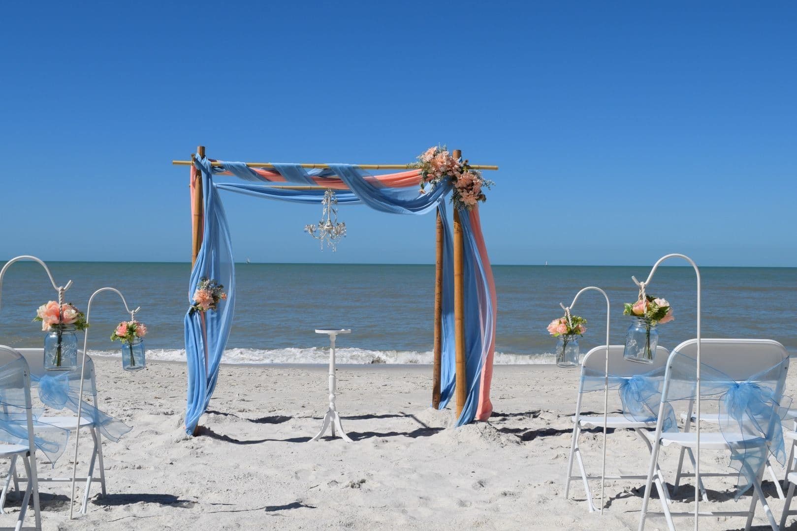 Beach wedding setup with an arch decorated in blue and orange fabric, chairs, and ocean backdrop.