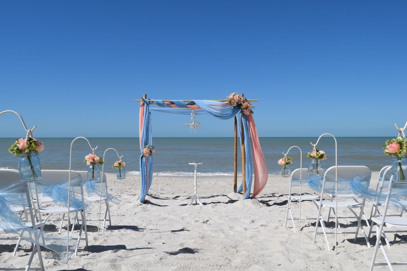Beach wedding setup: blue and coral draped arch, chairs, ocean backdrop.