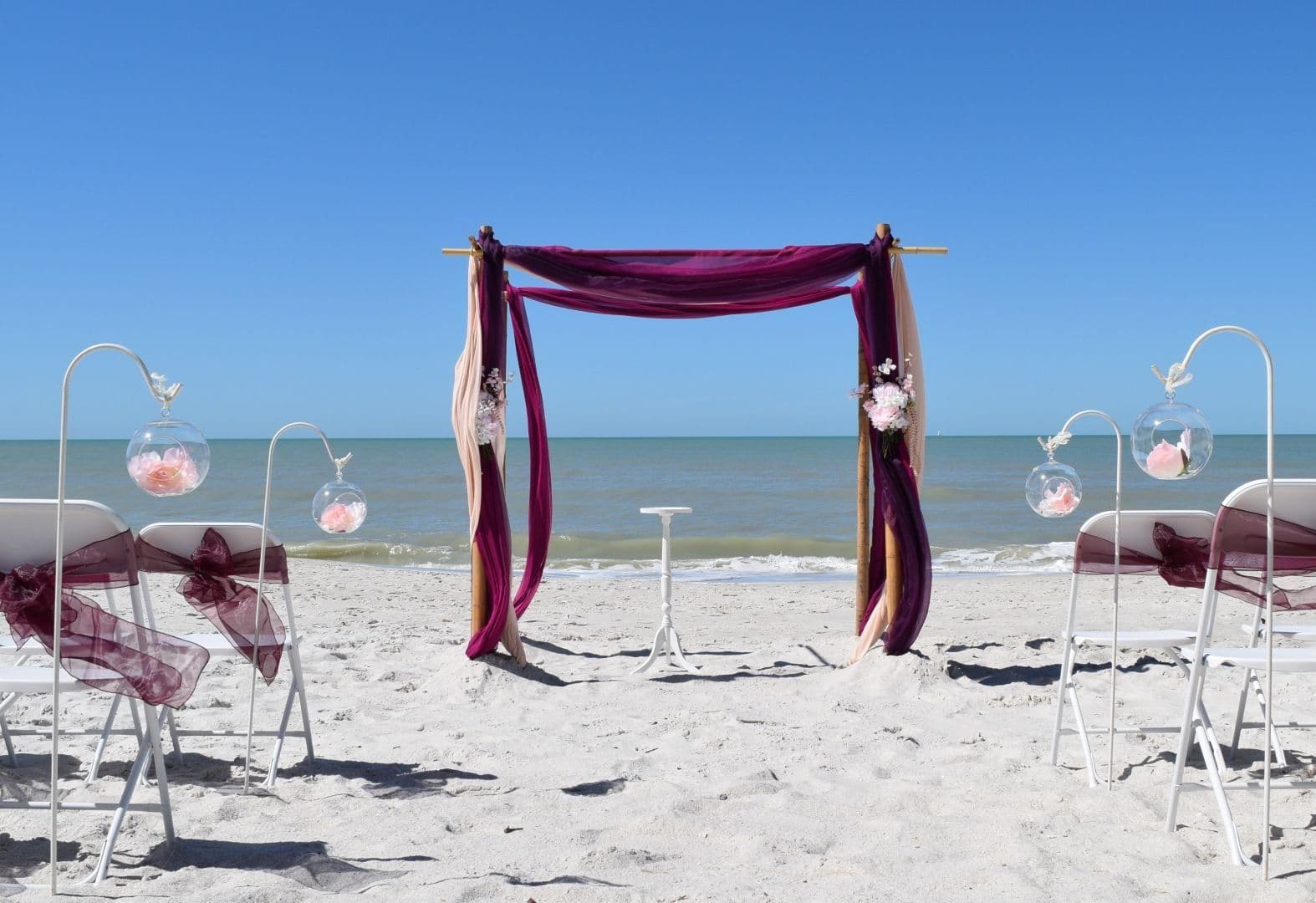 Beach wedding ceremony setup: purple draped arch, chairs with floral accents, clear sky and ocean backdrop.