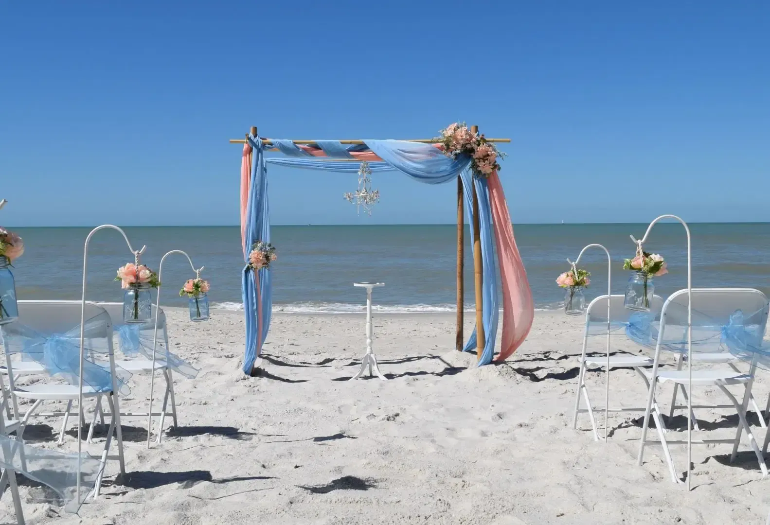 Beach wedding setup with draped arch, chairs, and ocean backdrop.