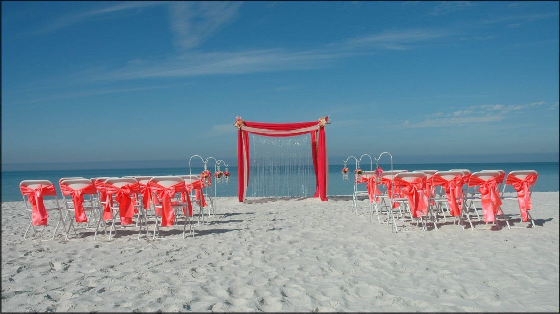 Beach wedding setup with red chairs and arch against a blue sky.