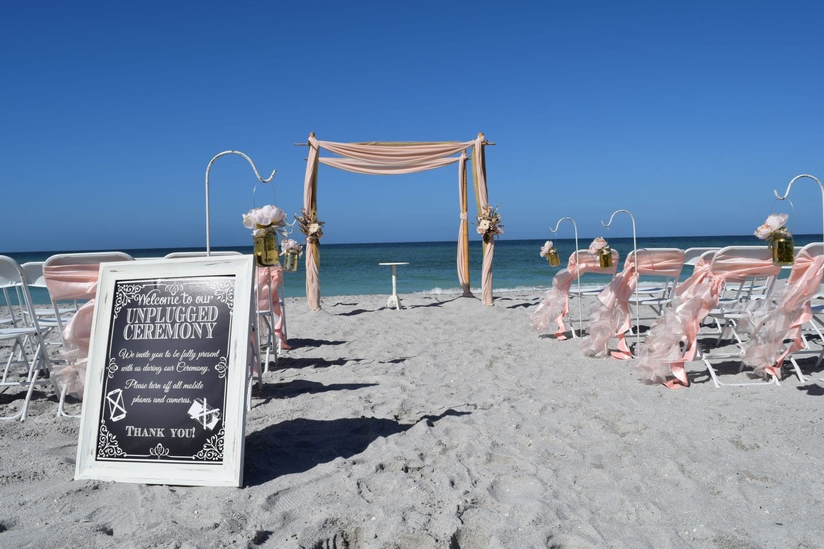 Beach wedding ceremony setup with white chairs, archway, and sign on a sunny day.