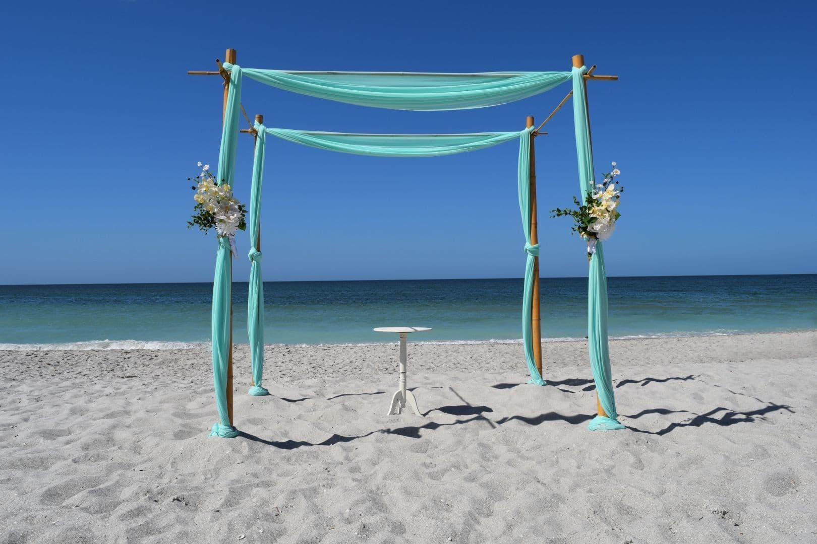 Wedding arch on a beach with turquoise fabric, flowers, and a small table, against a blue sky and ocean.