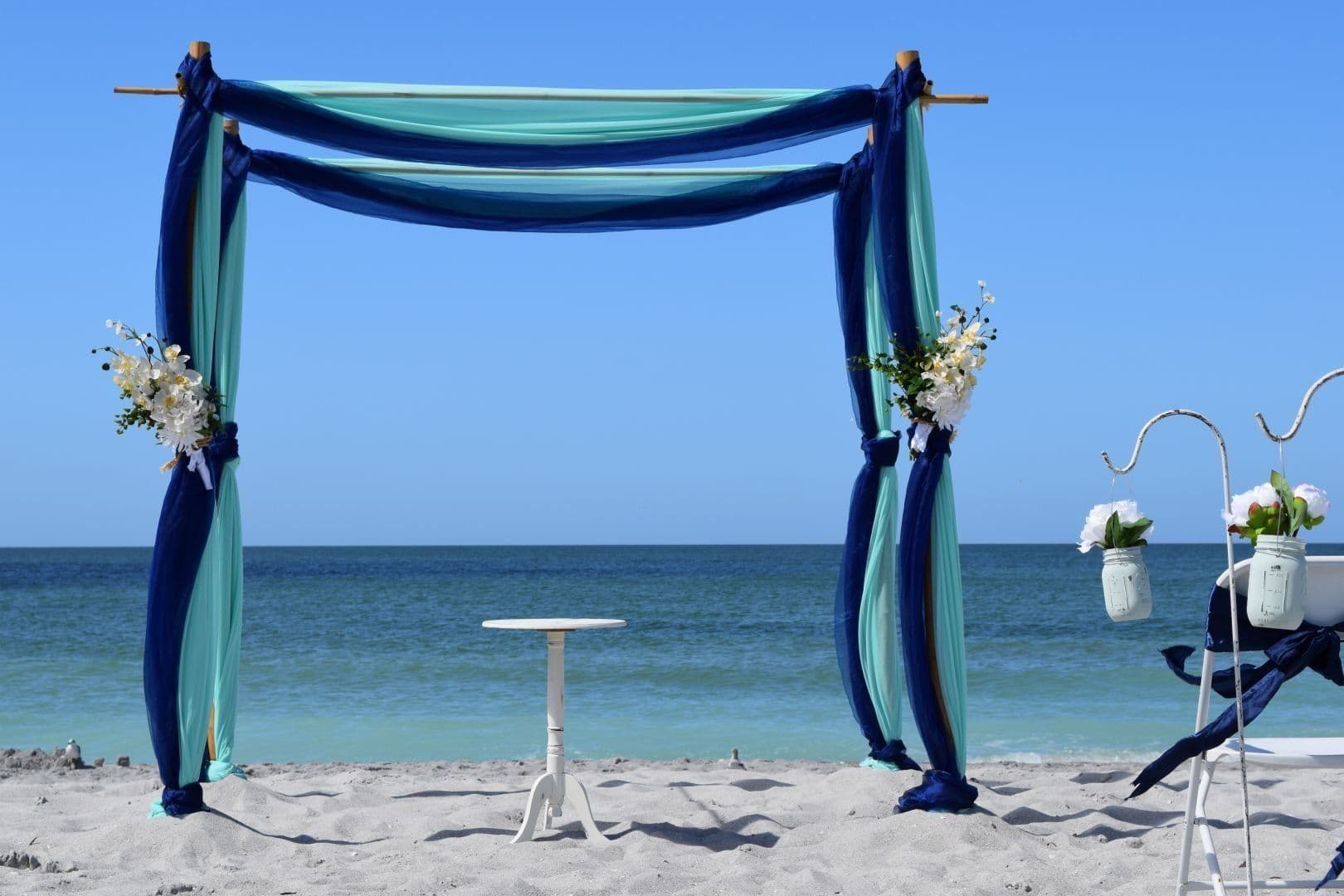 Beach wedding arch draped with teal and navy fabric; small table; ocean and blue sky in background.