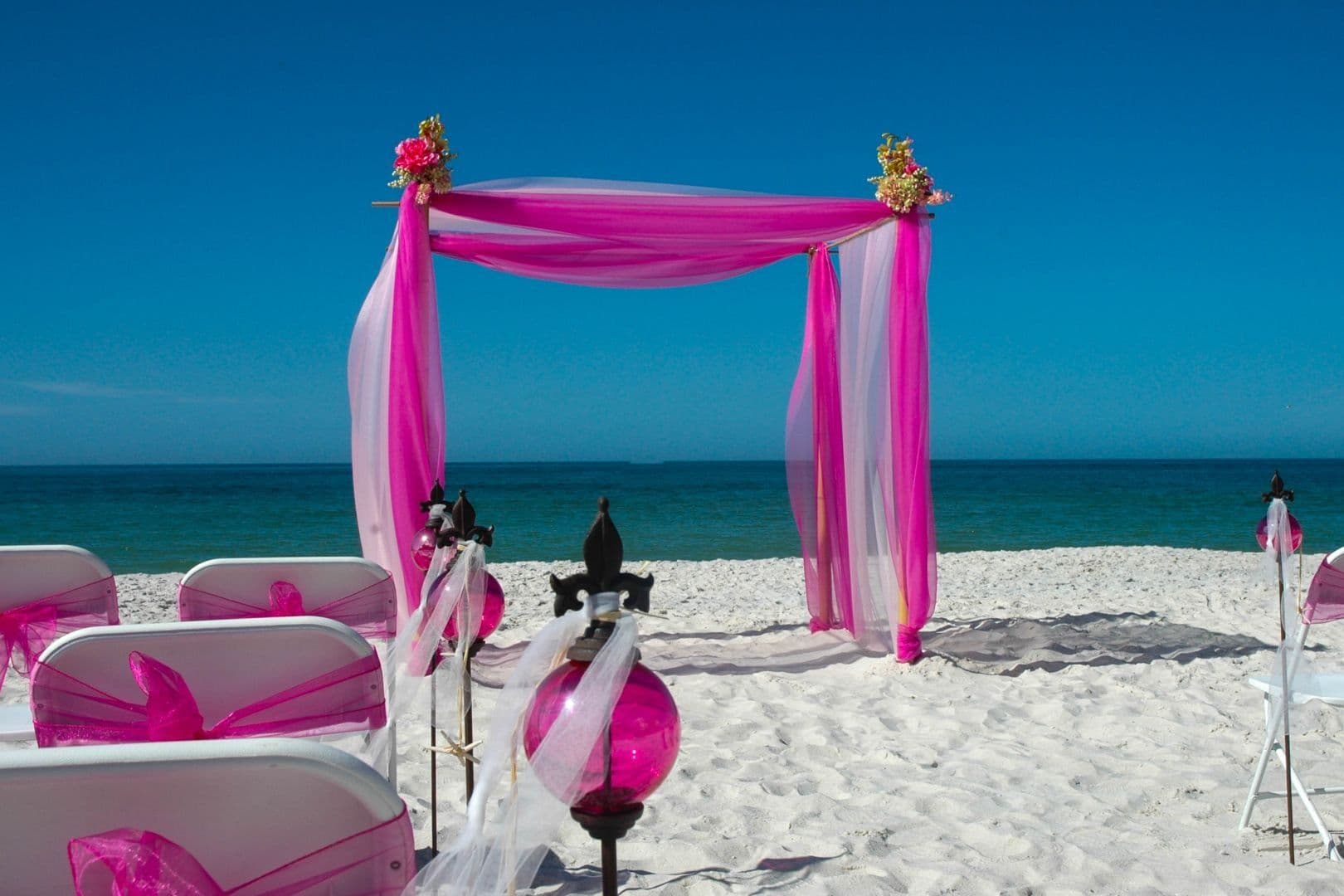 Beach wedding ceremony setup with pink and white decorations; blue sky and ocean.