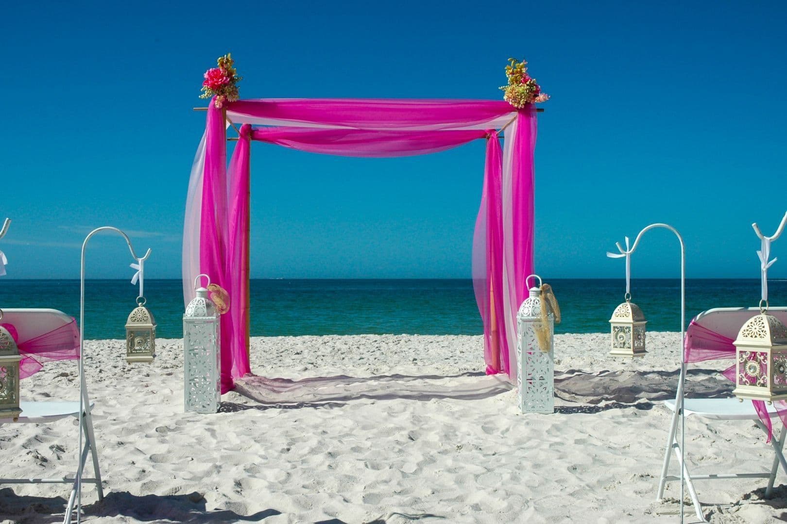 Beach wedding ceremony setup: Pink draped arch, white sand, blue ocean, lanterns, chairs, clear sky.