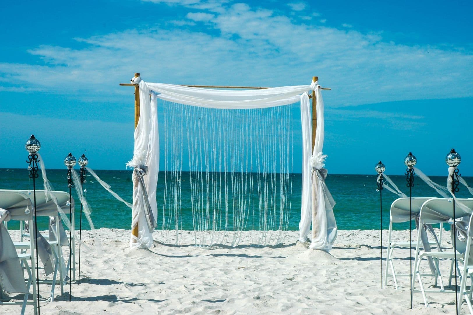 Beach wedding setup: white draped arch, chairs, and decorations on the sand facing the ocean.