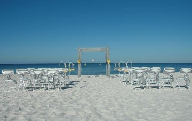 Beach wedding setup: white chairs on sand facing a decorated arch with the ocean in the background under a blue sky.