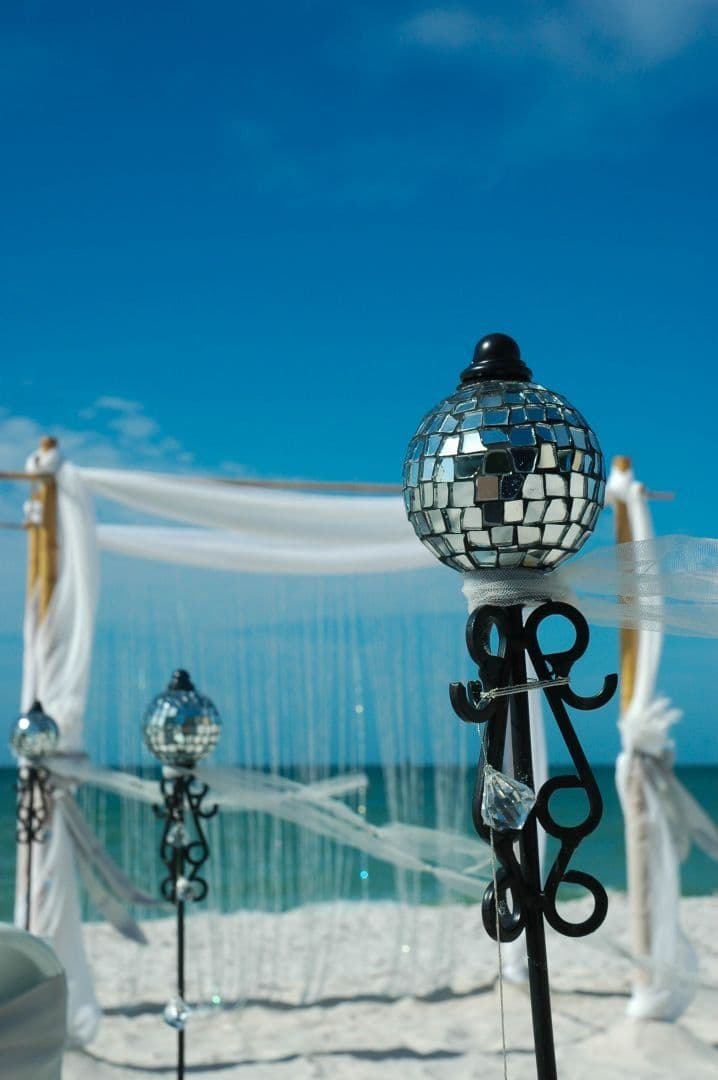 Beach wedding decor: disco ball atop black stand; white draping, blue sky, and sea.