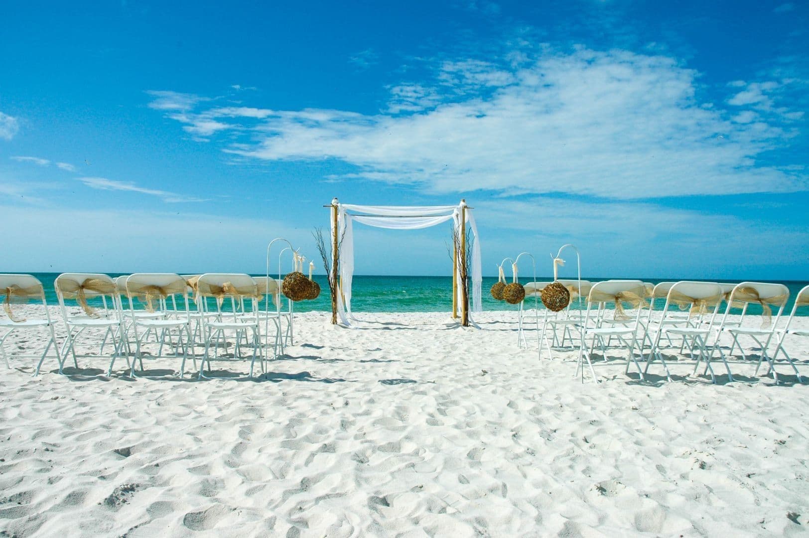 Beach wedding setup with chairs facing arch, white sand, turquoise water, blue sky.