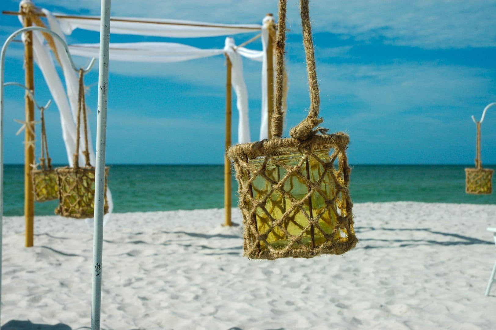 Beach wedding decor: Rope-wrapped lanterns hang from white poles, ocean and blue sky background.