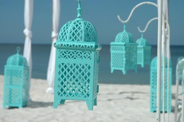 Teal lanterns hanging on a beach with white drapes, ocean, and sky background.