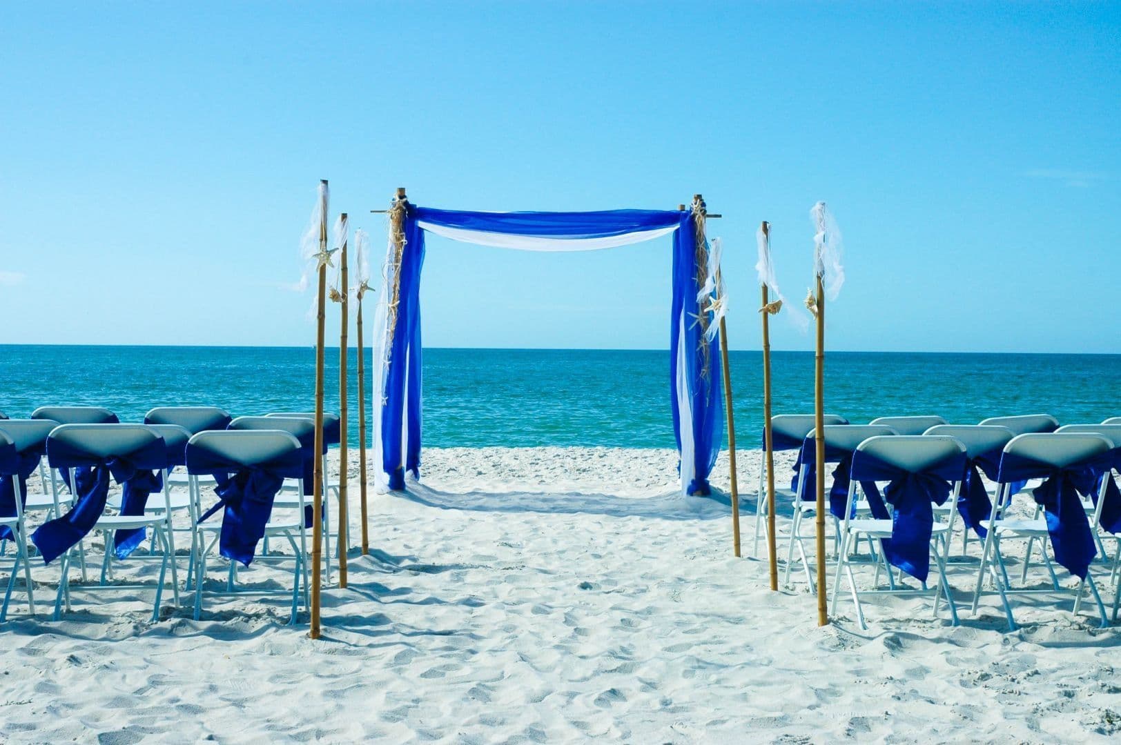 Beach wedding setup with blue accents; chairs face an arch draped in blue fabric, ocean in background.