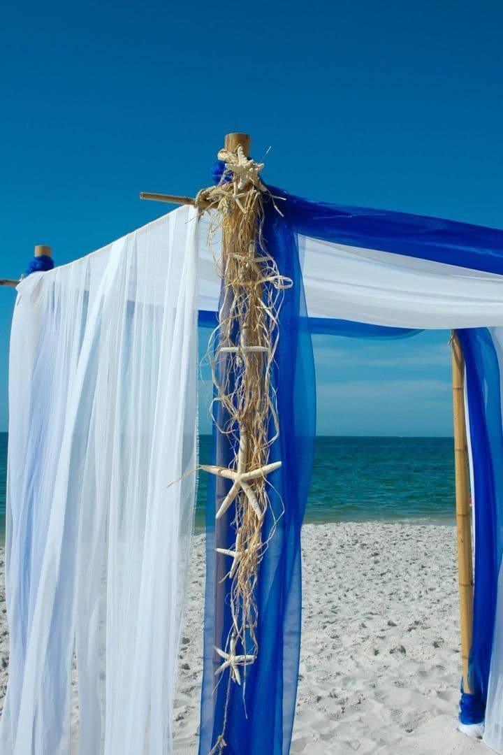 Beach wedding arch decorated with blue and white fabric, starfish, and natural garland.