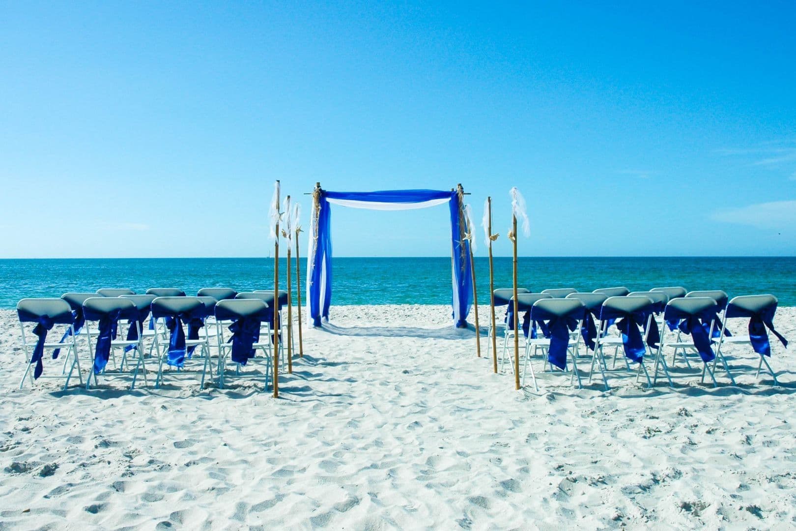 Beach wedding ceremony setup: blue arch, chairs, and ocean view on a sunny day.