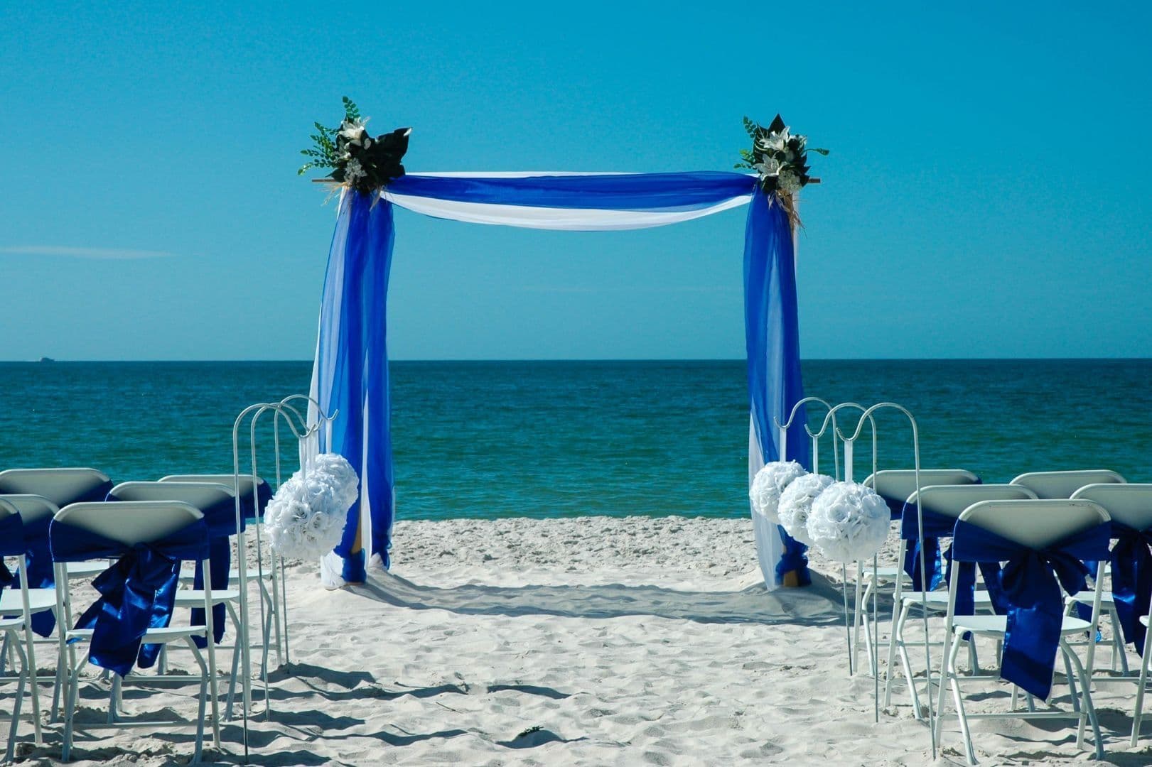 Beach wedding setup with blue arch and white chairs.