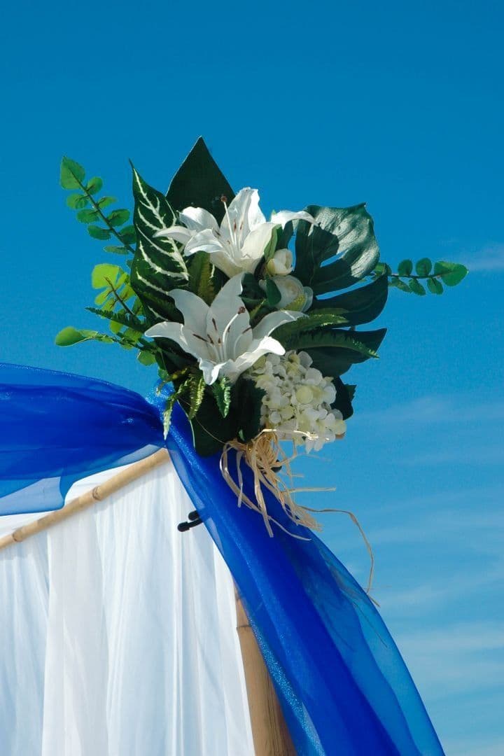Floral arrangement atop draped wedding arch against a clear blue sky.