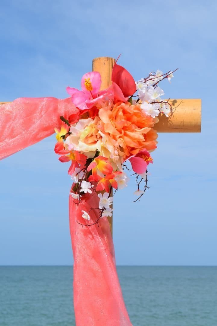Wooden cross decorated with flowers and coral fabric, against a blue sky and ocean.