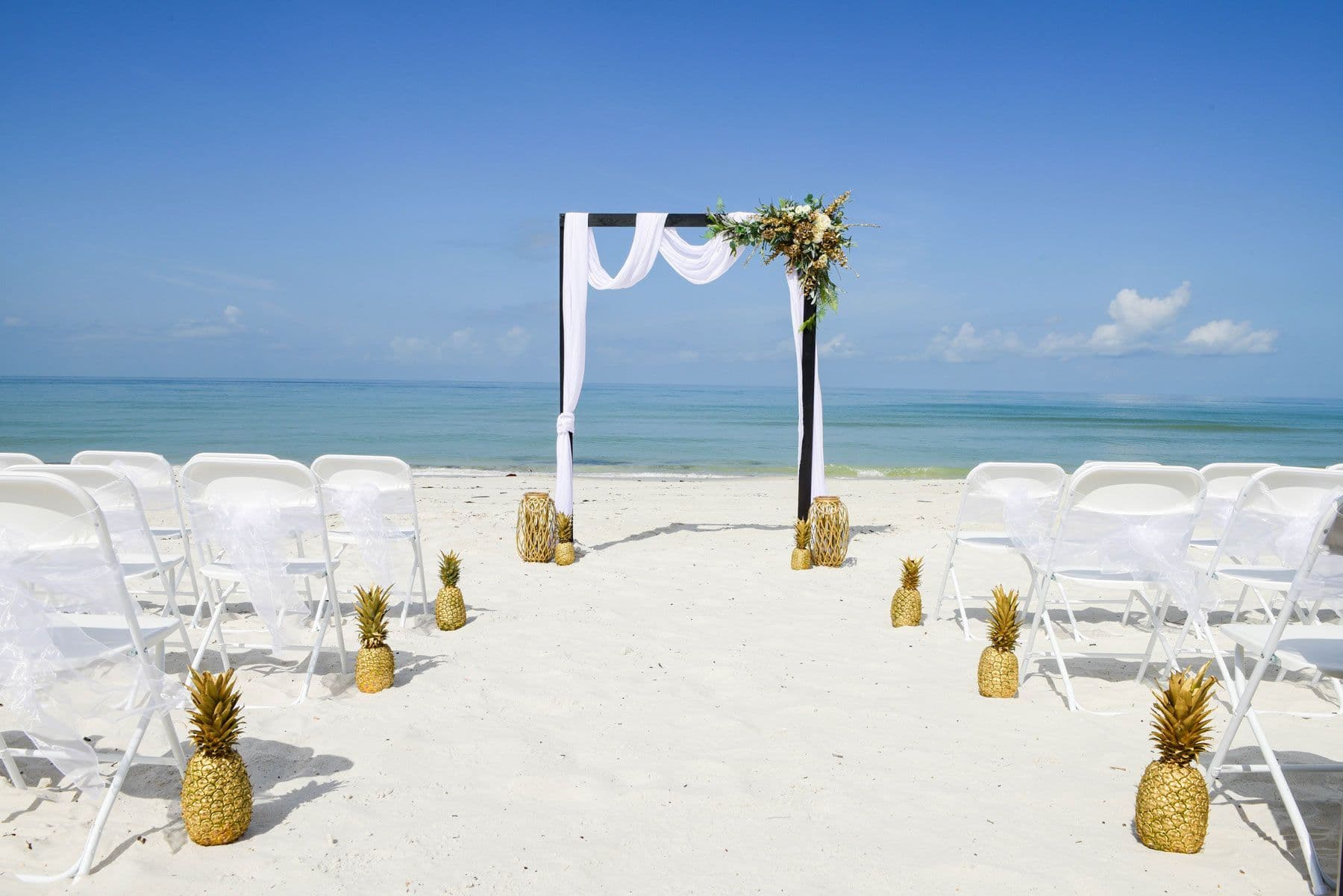 Beach wedding setup with white chairs, archway, and pineapple decorations on the sand.