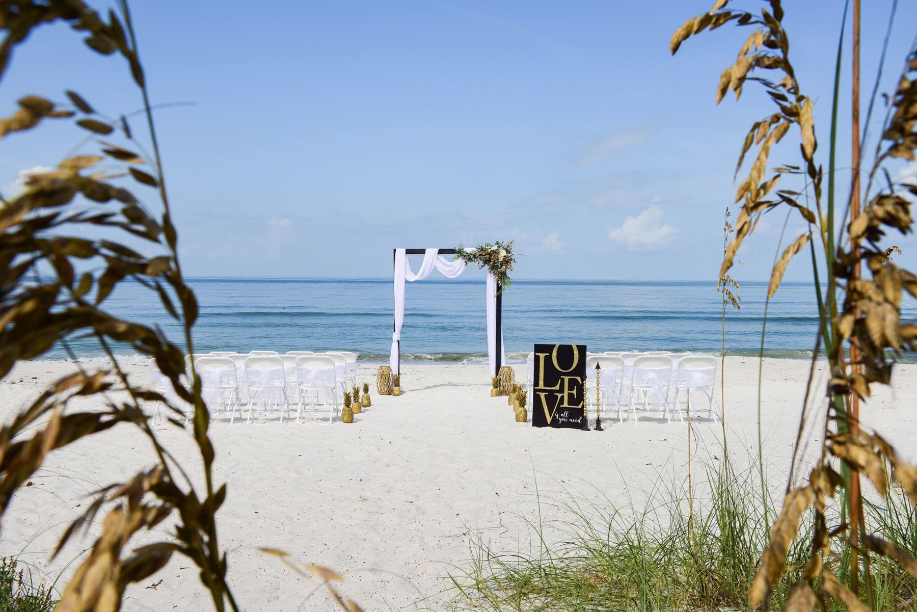 Beach wedding setup with chairs, arch, and “LOVE” sign, ocean backdrop.