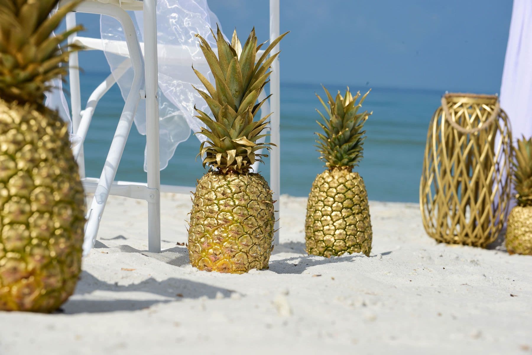 Golden pineapples on a sandy beach, white chairs and a blue sea in the background.