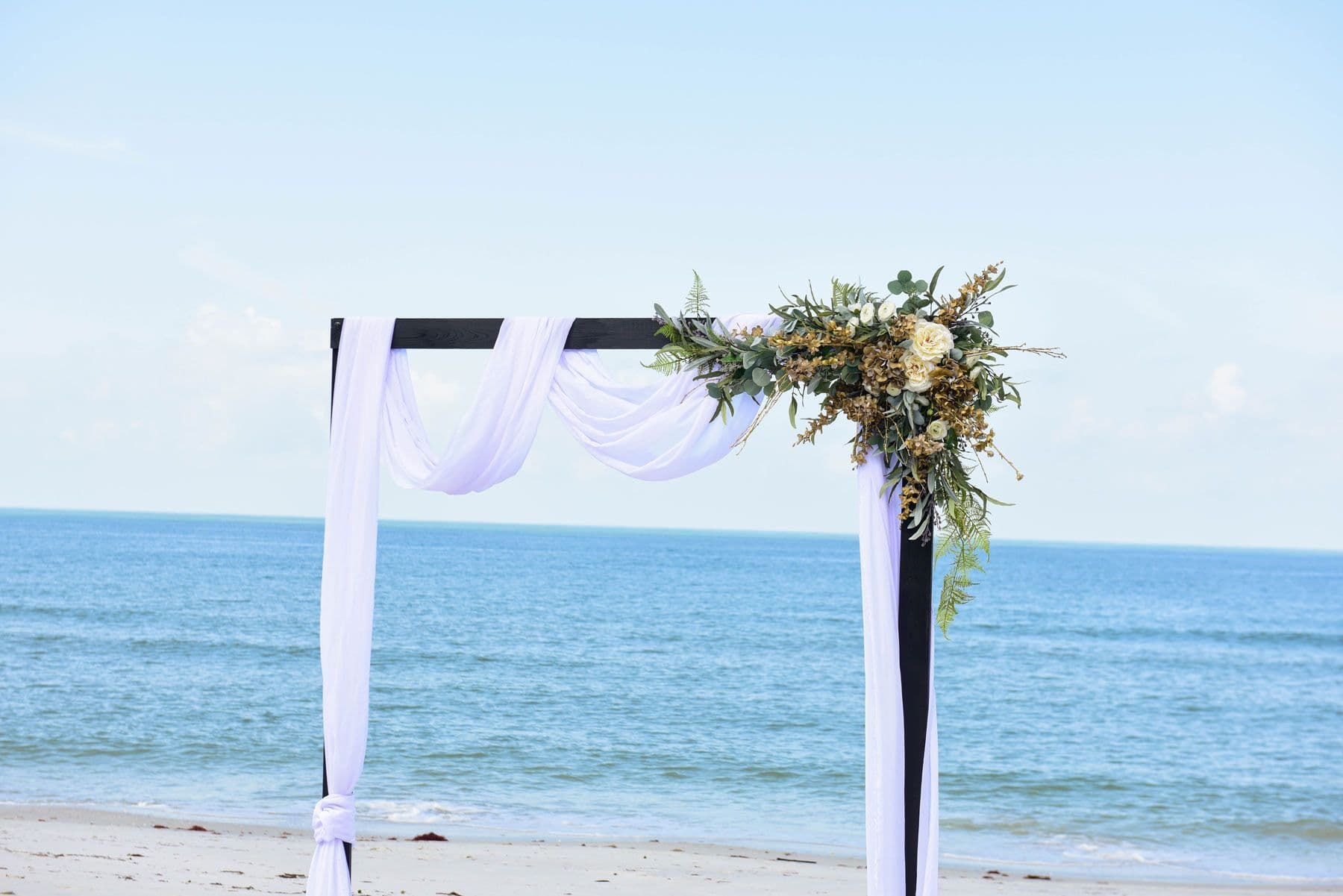 Wedding arch on a beach with white fabric and floral arrangement, ocean in background.