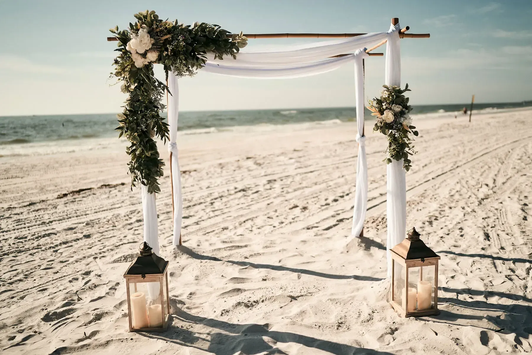 Wedding arch decorated with flowers and fabric on a beach, flanked by lanterns, ocean in background.