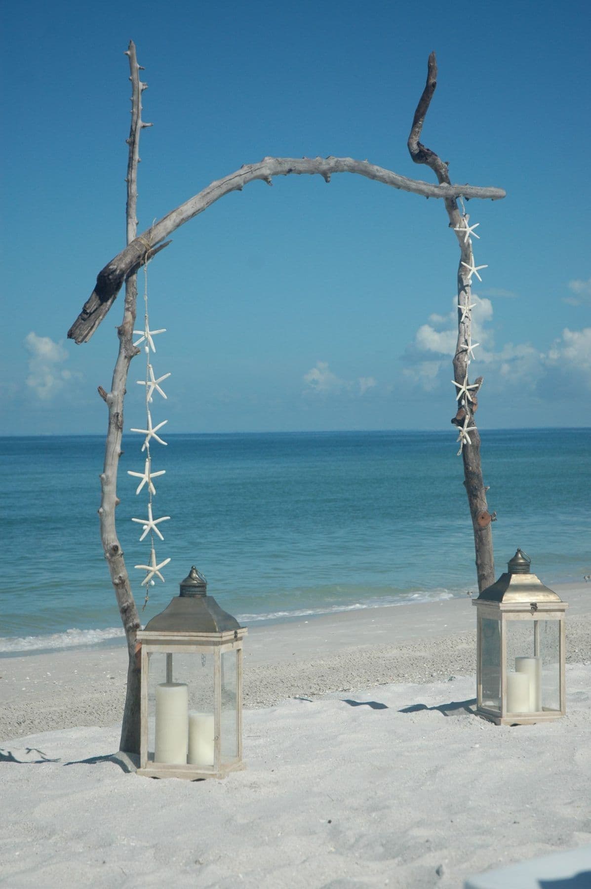 Driftwood arch with starfish and lanterns on a sandy beach, ocean in background.