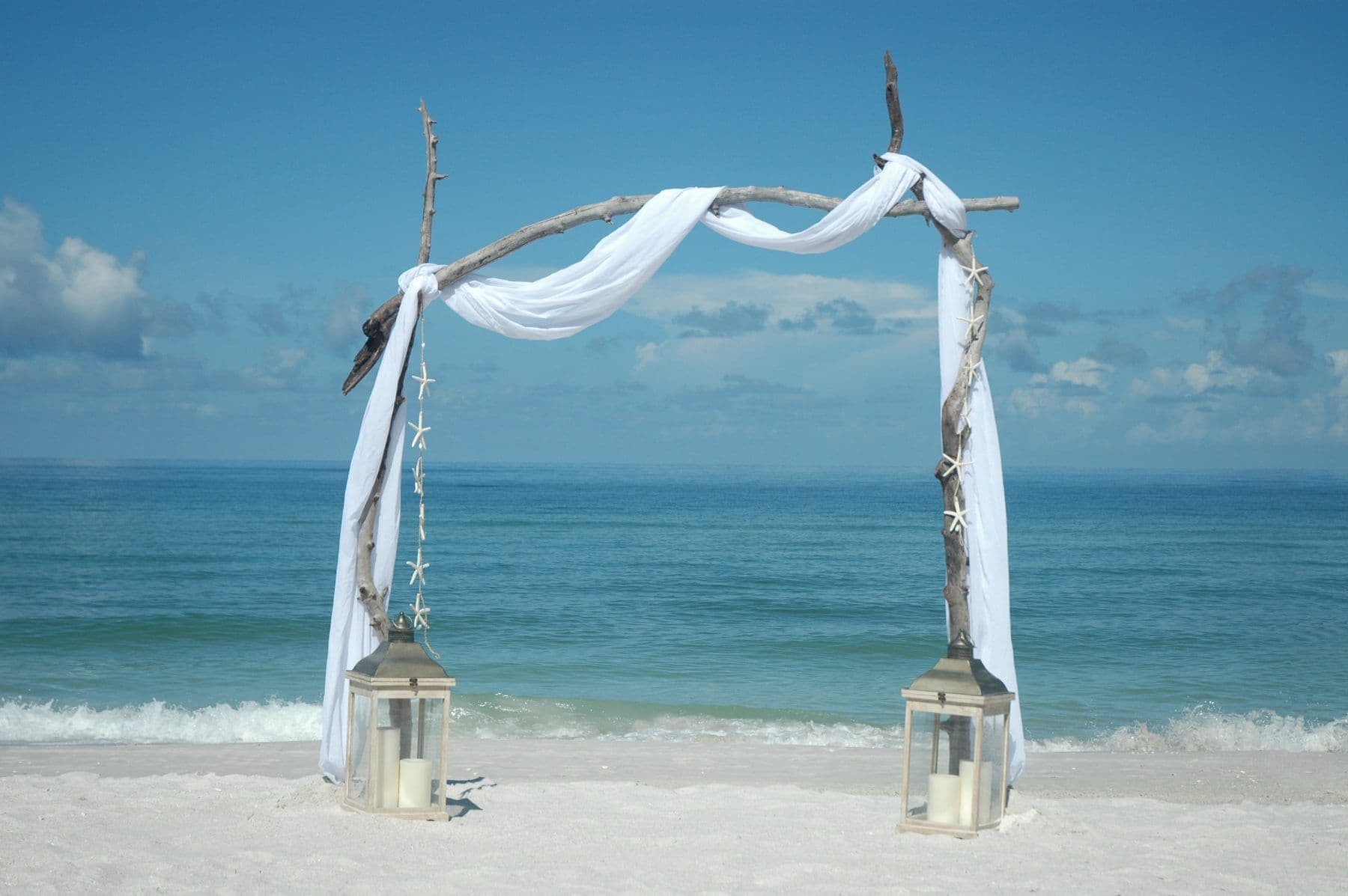 Beach wedding arch with white fabric, lanterns, and ocean backdrop.