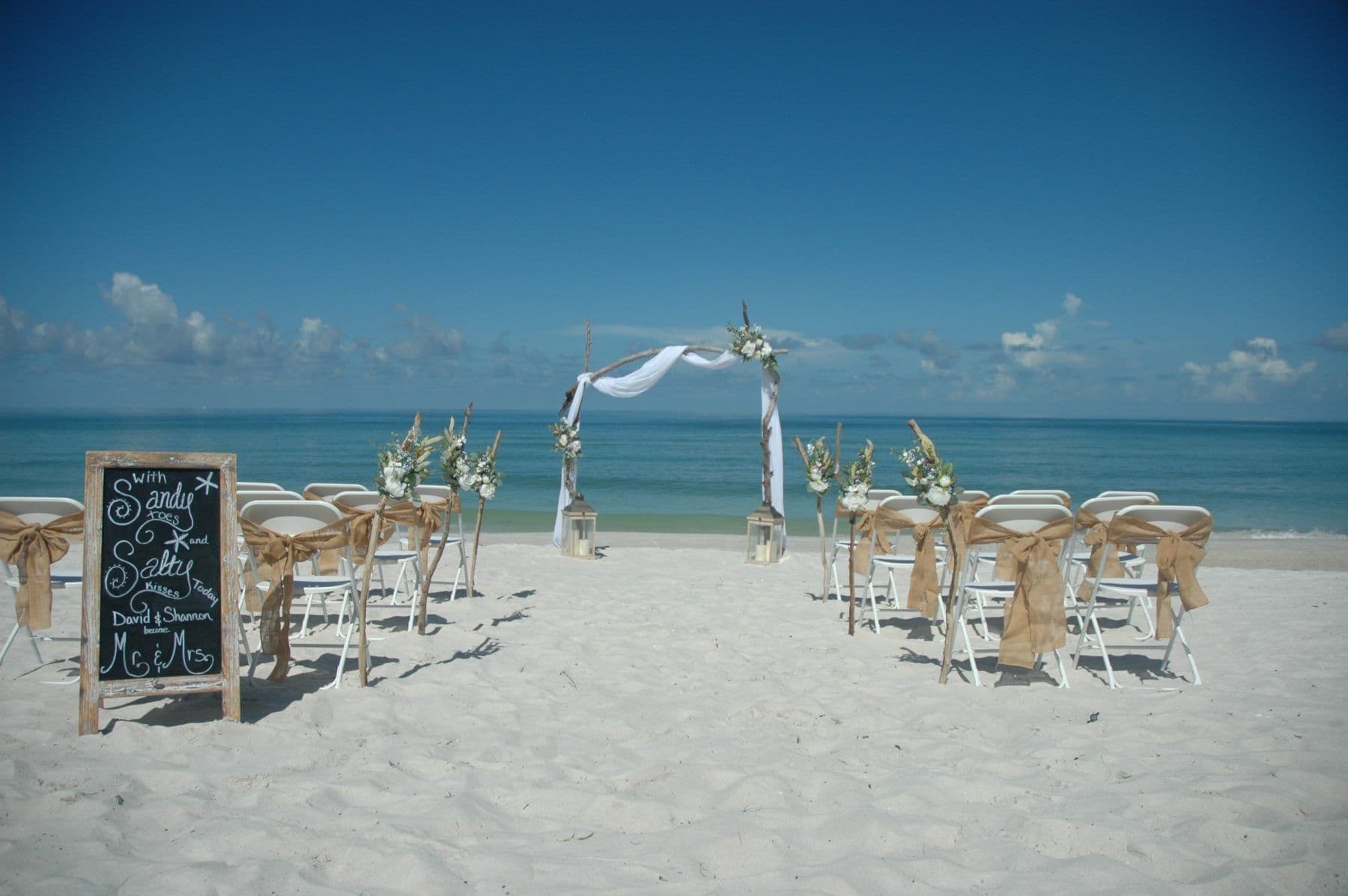 Beach wedding setup: white chairs with burlap accents face floral arch on sandy shore, blue sky and ocean backdrop.