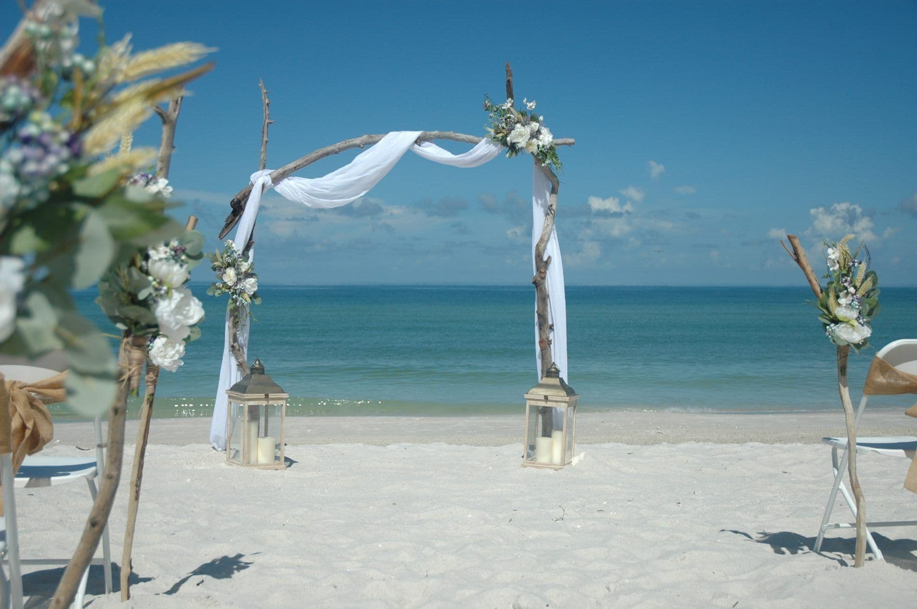 Beach wedding arch with white fabric, flowers, and lanterns against the ocean.