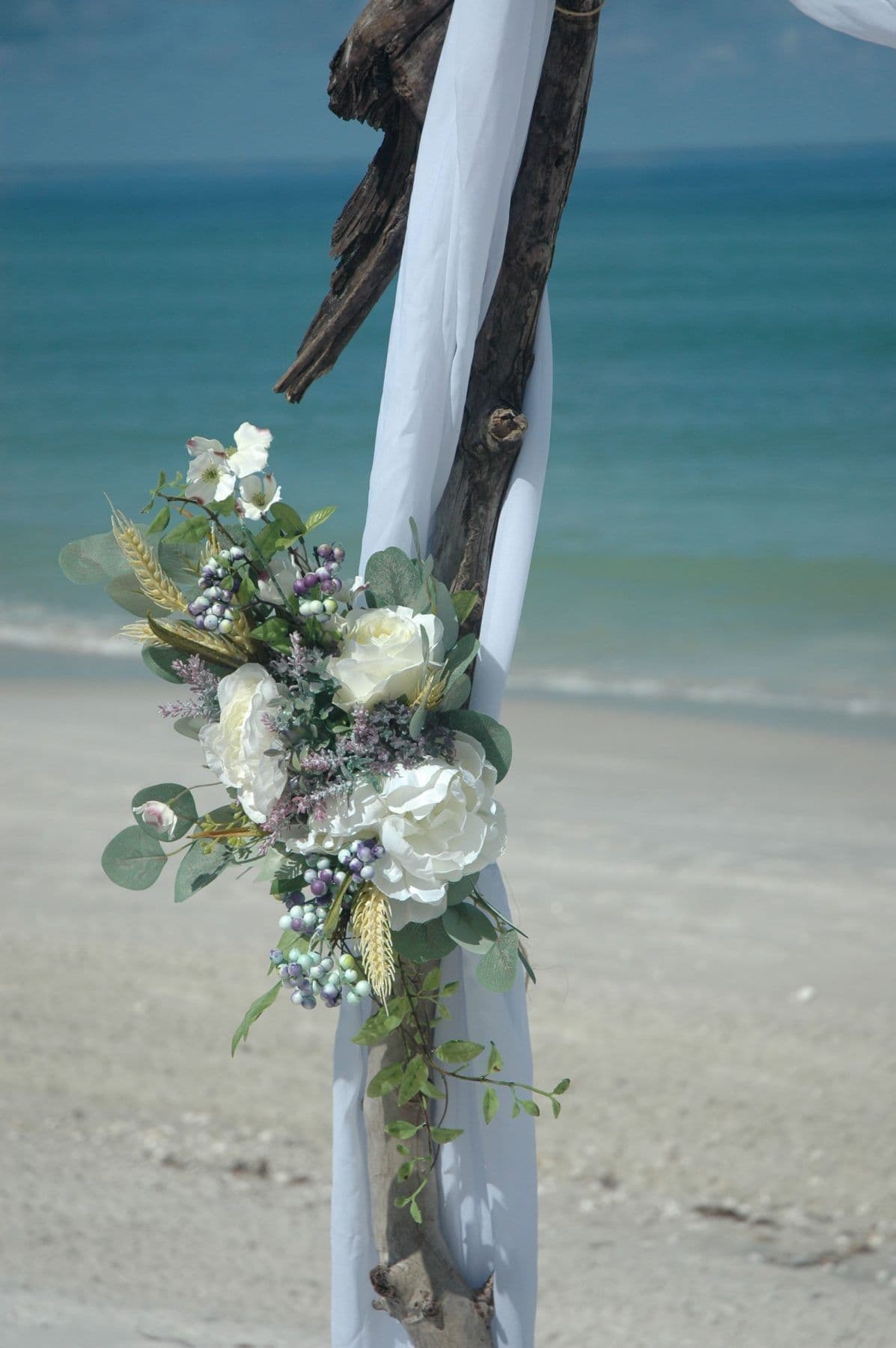 Beach wedding arch with white flowers and sheer fabric, driftwood on a sandy beach.