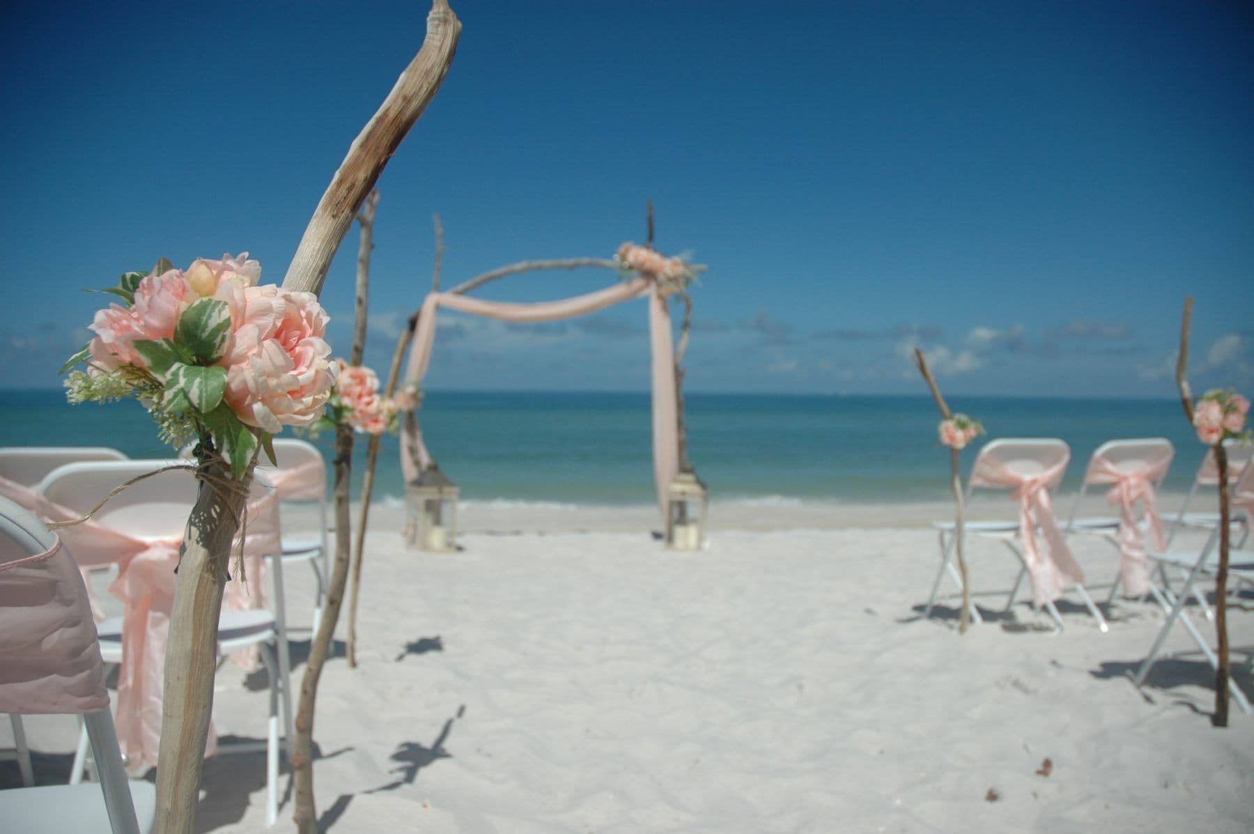 Beach wedding setup with arch, chairs, and pink decorations on white sand, blue sky and ocean.