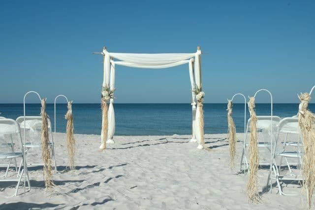 Wedding setup on a white sandy beach with chairs, arch, and ocean view on a sunny day.