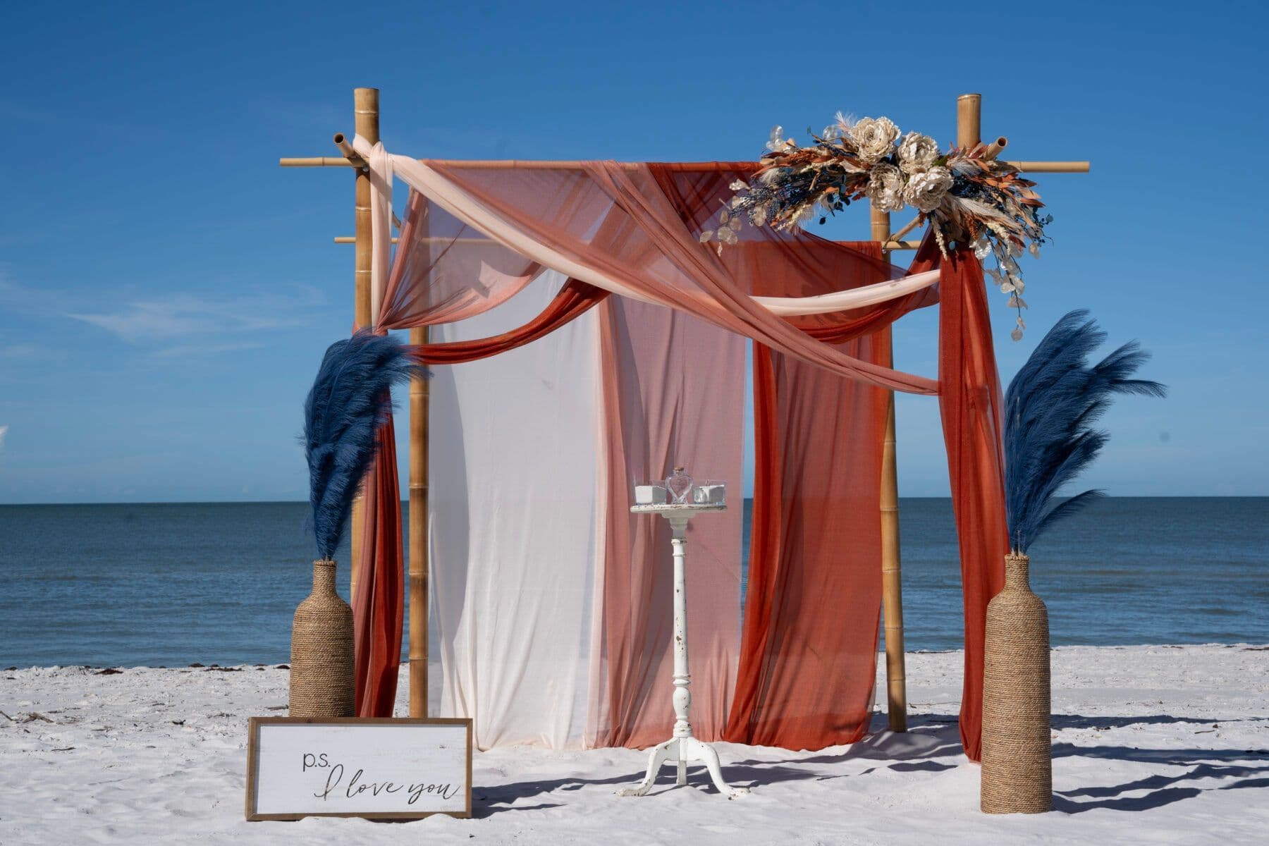 Beach wedding arch with red and white draped fabric, bamboo frame, and blue plumes.