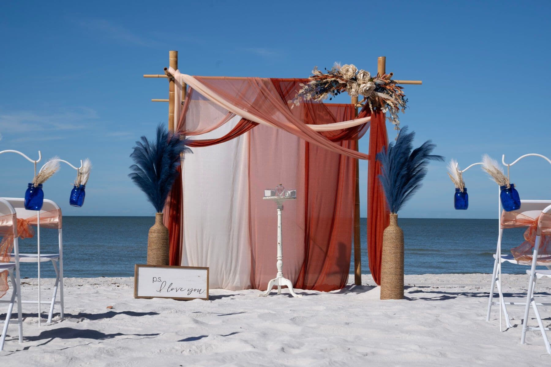 Beach wedding setup with draped fabric backdrop, floral accents, and two rows of chairs.