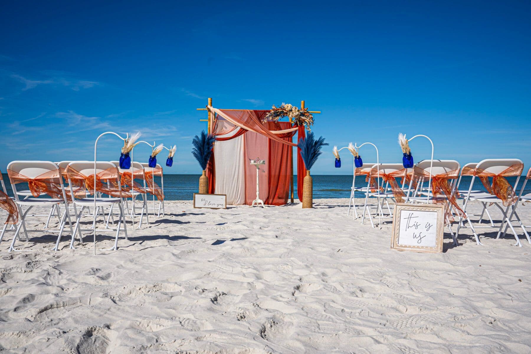 Beach wedding setup with chairs, an arch, and decorations against a blue sky.